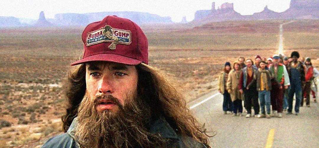 A man with a long beard and a red Bubba Gump cap stands on a deserted road, looking thoughtful. A group of people follows him in the distance, with a vast, rocky desert landscape in the background.