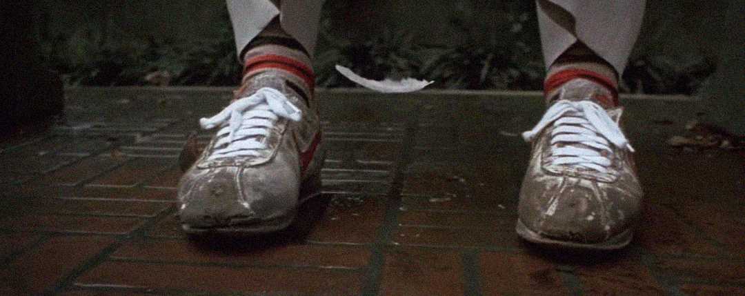 A white feather floats down in front of worn, mud-stained sneakers with white laces and red stripes, worn by someone standing on wet brick pavement. The person is also wearing white pants and striped socks.