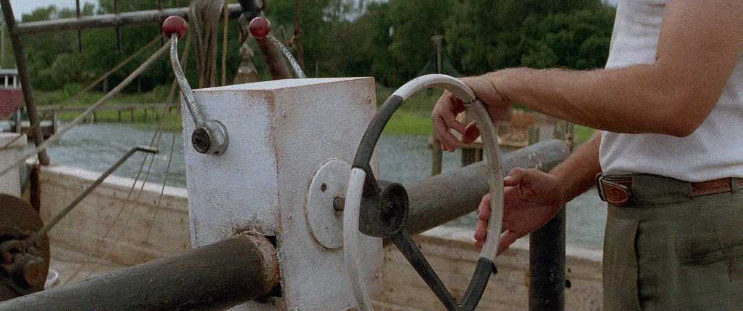 A person wearing a white shirt and green pants steers a boat using a white and black metal wheel mounted on a weathered metal console near water, with greenery visible in the background.