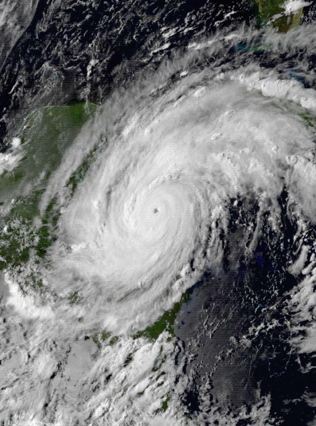 A satellite image shows a large, swirling hurricane with a well-defined eye centered over land and water, surrounded by thick clouds covering parts of the coastline and ocean.