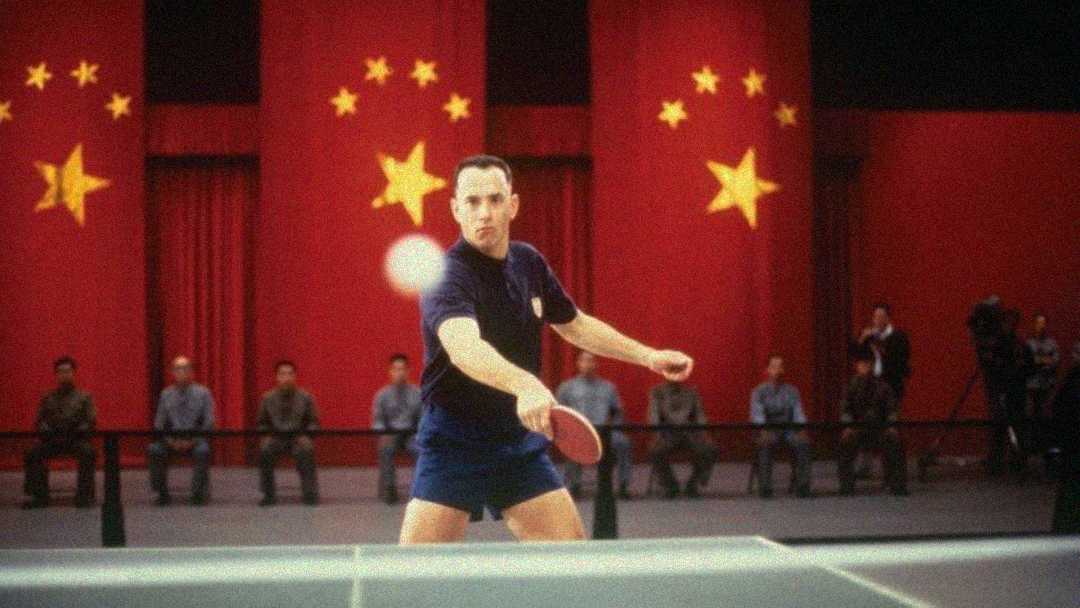 A man in blue athletic wear plays table tennis in front of a row of people seated and standing, with large red curtains and five golden Chinese flags displayed in the background.