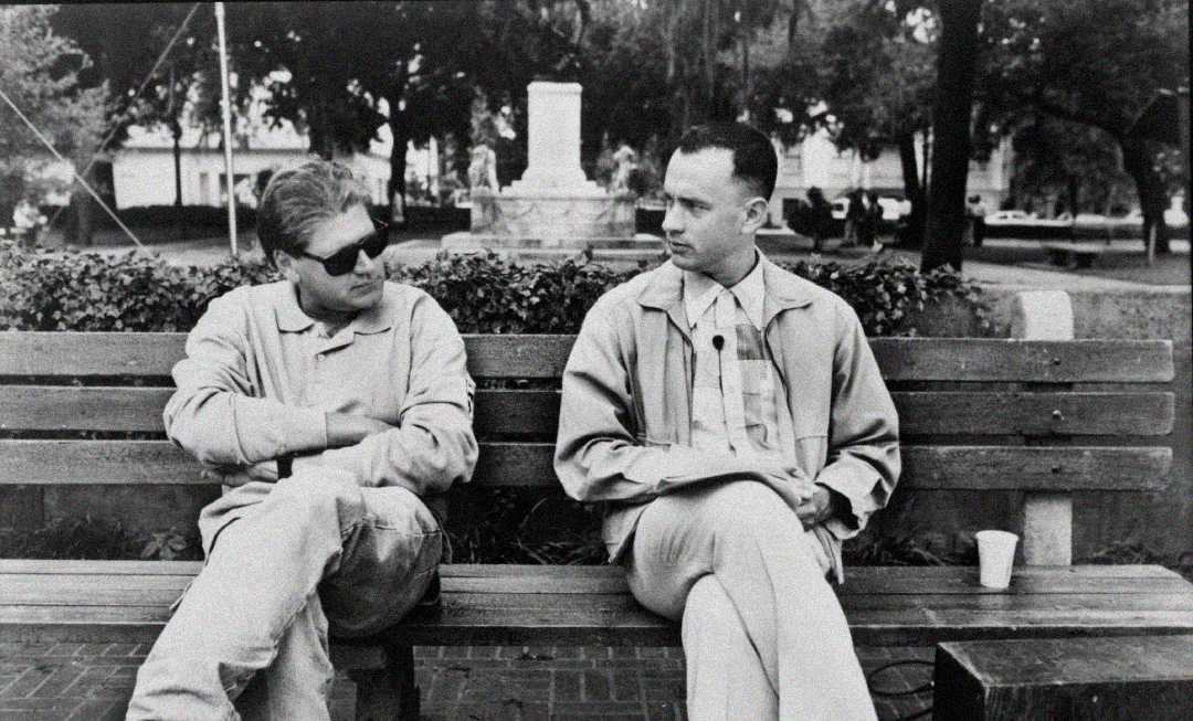 Two men sit on a park bench, facing each other. One wears sunglasses and casual clothes, while the other wears a jacket and has his legs crossed. Trees and a monument appear in the background. The image is black and white.