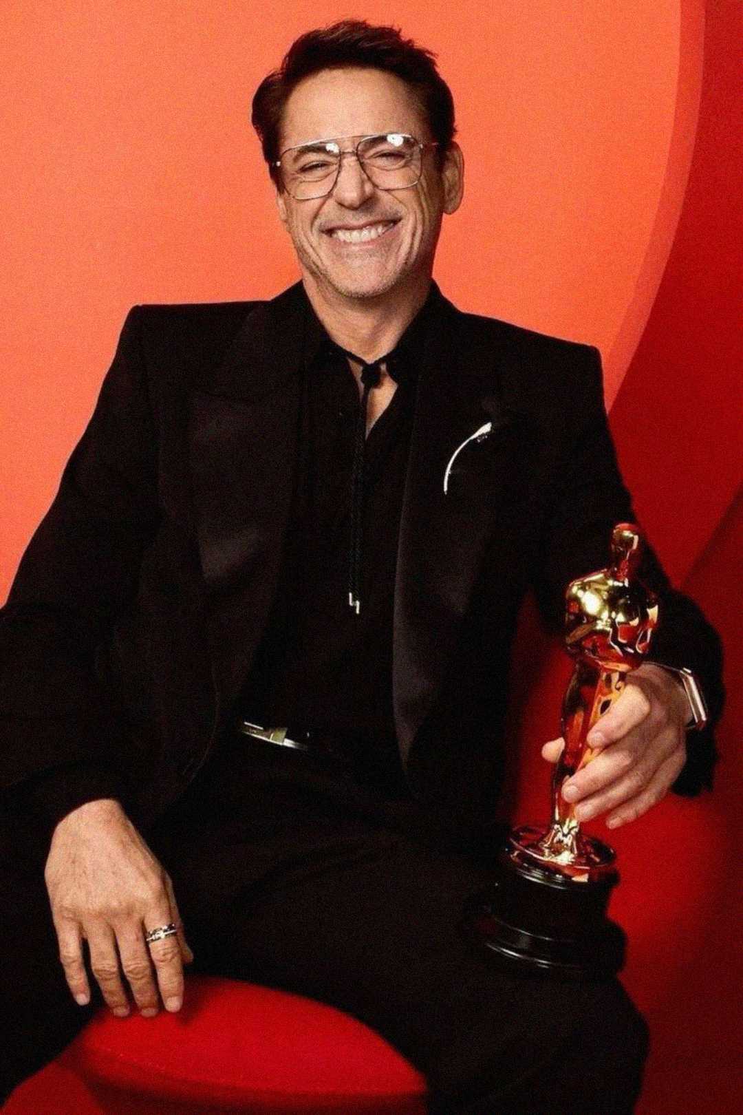 A man in a black suit and glasses smiles while seated on a red chair, holding a gold Oscar statuette against a red background.