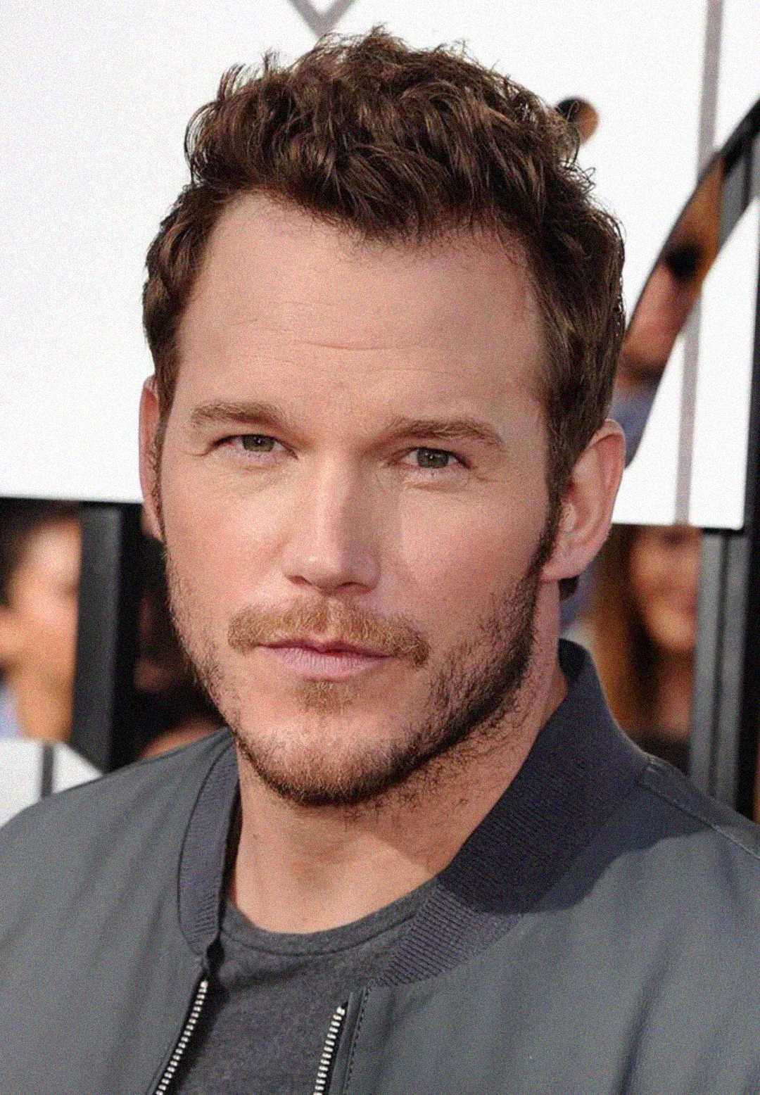 A man with short, wavy brown hair and light stubble wears a gray jacket over a t-shirt, looking directly at the camera with a neutral expression. The background is blurred.