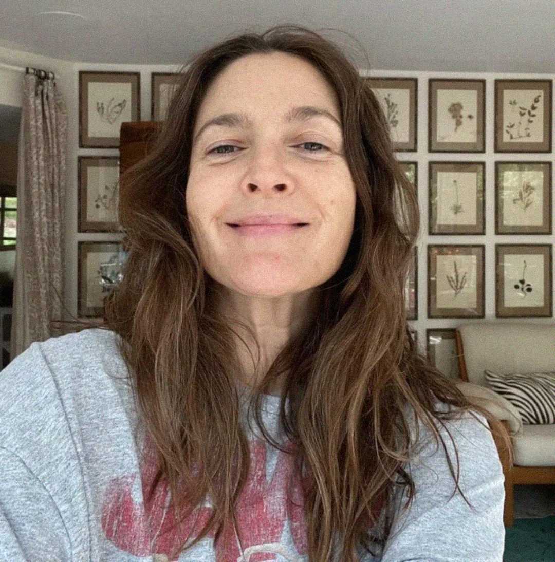 A woman with long brown hair and a natural look smiles gently at the camera in a cozy room with framed botanical prints covering the wall behind her. She is wearing a light gray top.