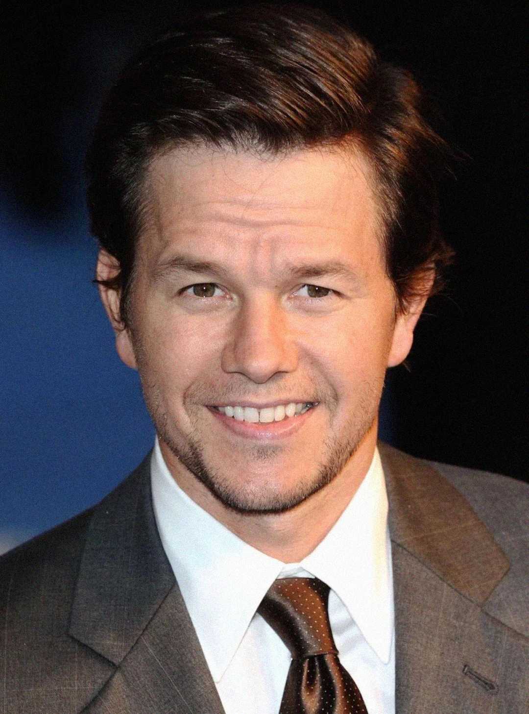 A man with short brown hair and light stubble smiles at the camera. He is wearing a gray suit, white shirt, and brown tie, with a dark background behind him.