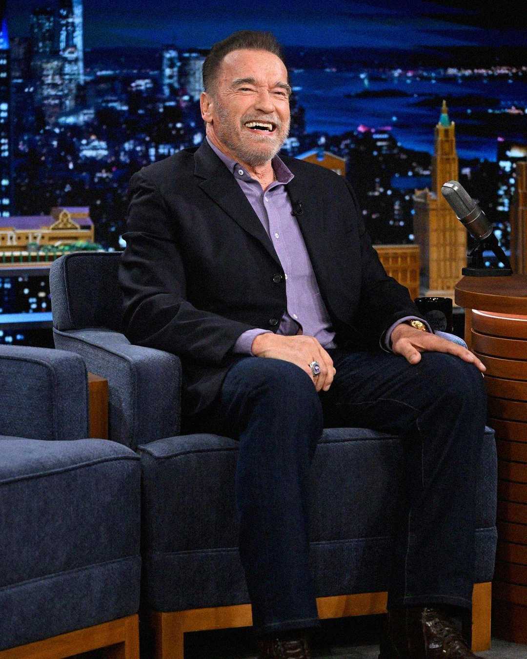 A man in a dark suit and lavender shirt sits smiling and laughing on a talk show set, with a cityscape background and a microphone on the desk beside him.