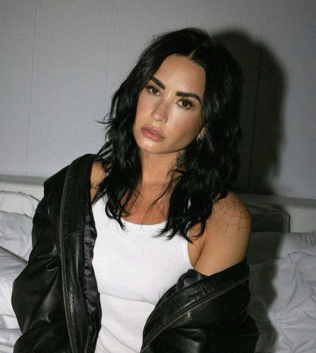 A person with long, wavy black hair sits on a bed, wearing a white tank top and a black leather jacket draped over their shoulders, looking directly at the camera. The background is simple and softly lit.