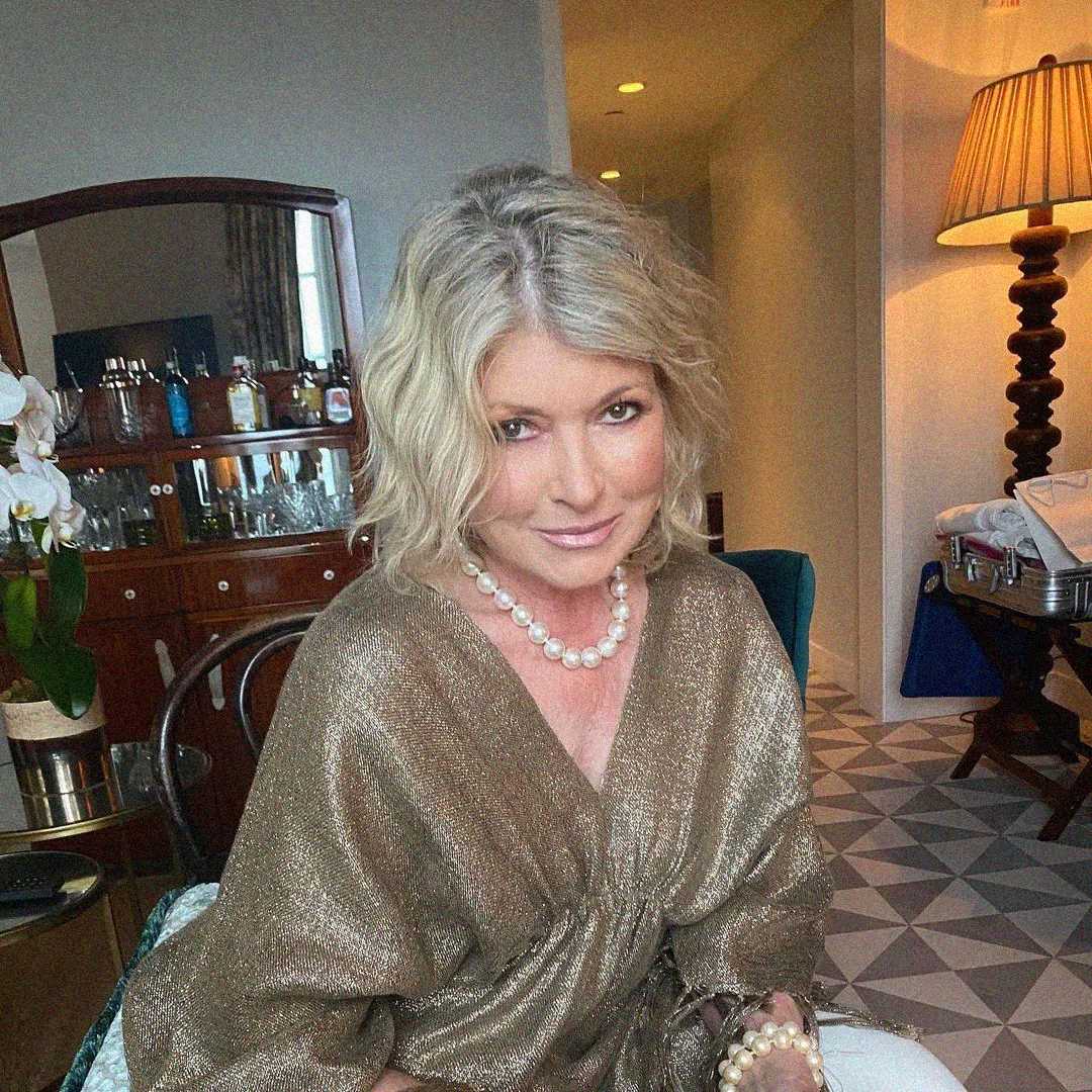 A woman with wavy blonde hair wearing a gold shimmery top and a pearl necklace sits in a stylish room with a bar cabinet, orchids, and a lamp in the background. She is smiling softly at the camera.