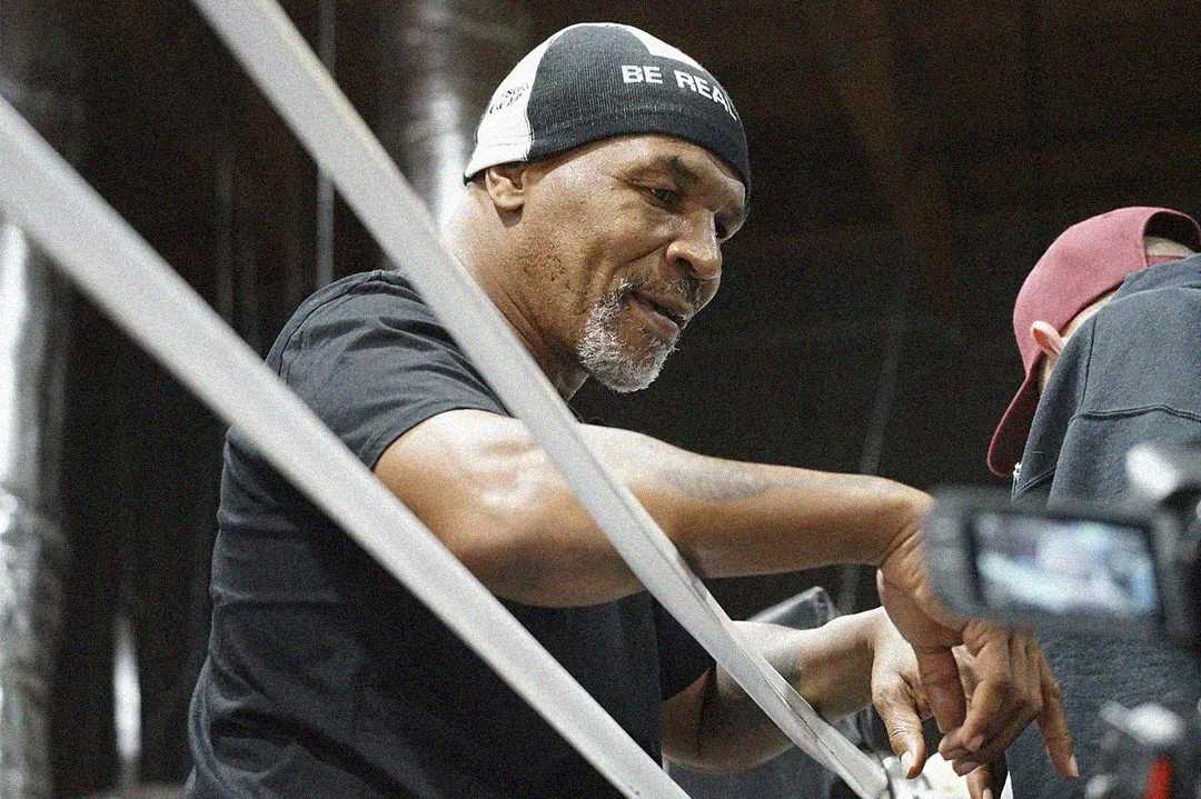 A man wearing a black beanie that says "BE REAL" and a black t-shirt is wrapping his hands with boxing tape in a gym setting, preparing for a workout. Another person stands nearby, partially out of view.