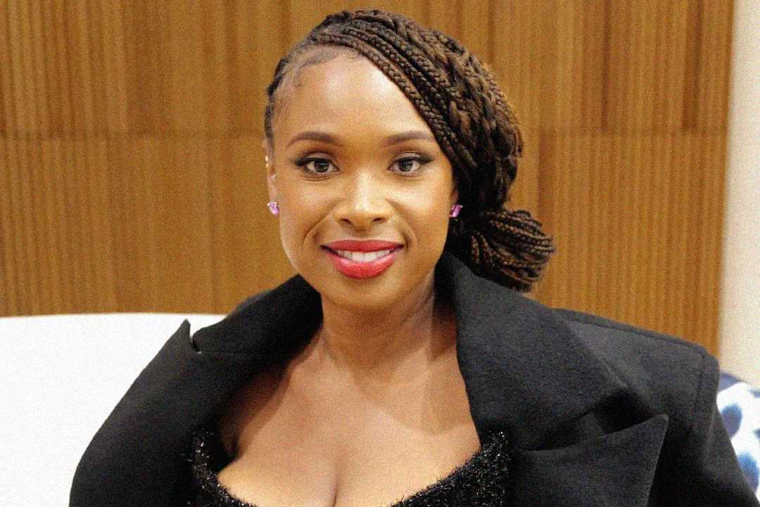 A woman with braided hair styled to one side, wearing a black off-shoulder outfit, smiles at the camera. She has bright red lipstick and subtle earrings, with a neutral background behind her.