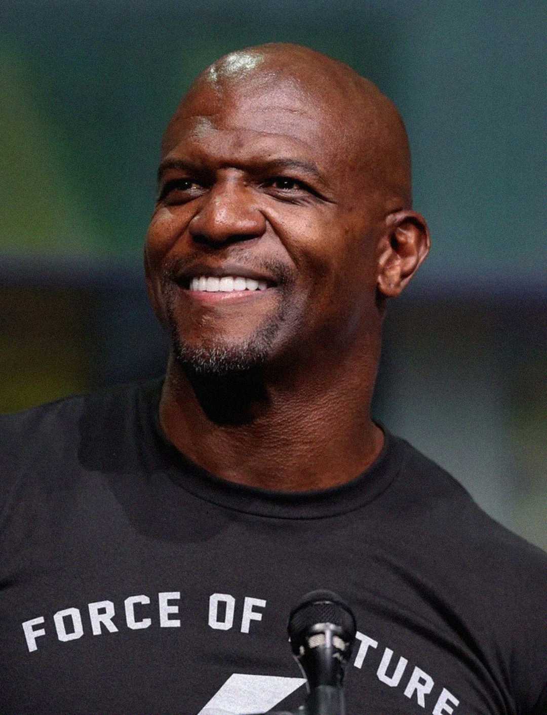 A smiling bald man with a goatee wearing a black "Force of Nature" T-shirt stands in front of a microphone, with a blurred background behind him.