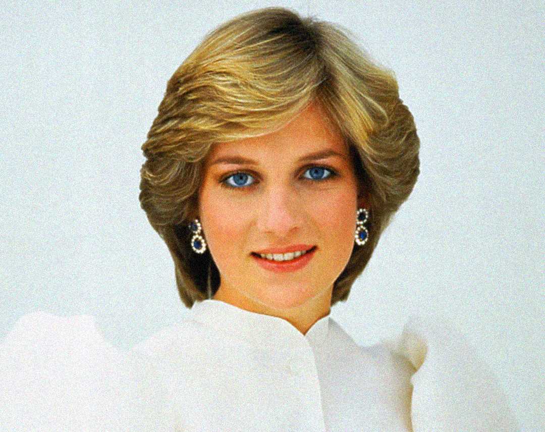 A woman with short, light brown hair and blue eyes smiles softly. She is wearing a white blouse with puffed shoulders and elegant drop earrings against a plain light background.