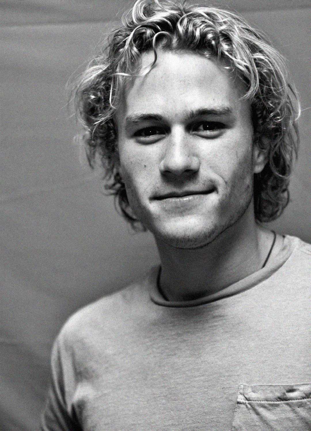 A black-and-white portrait of a young man with wavy blond hair, wearing a light-colored T-shirt and looking into the camera with a slight smile. The background is plain and softly lit.