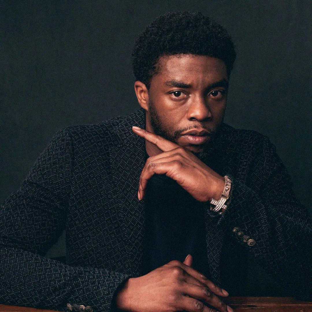 A man with short curly hair and a trimmed beard sits at a table, resting his chin on one hand and looking thoughtfully at the camera. He wears a dark textured blazer and a watch, with a dark background behind him.