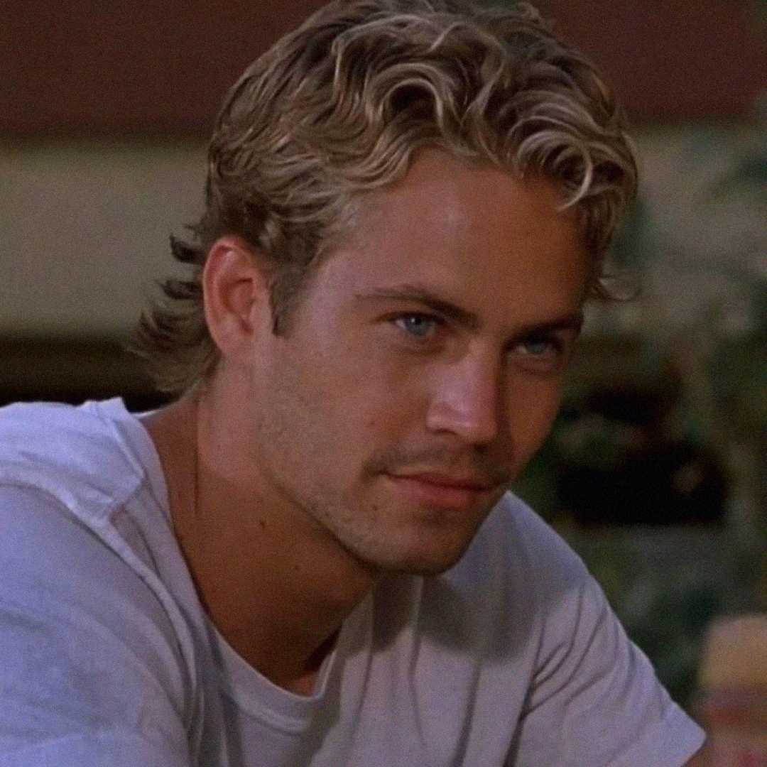 A young man with wavy, blonde hair and blue eyes looks slightly to the side, wearing a light-colored shirt and sitting outdoors in soft lighting.