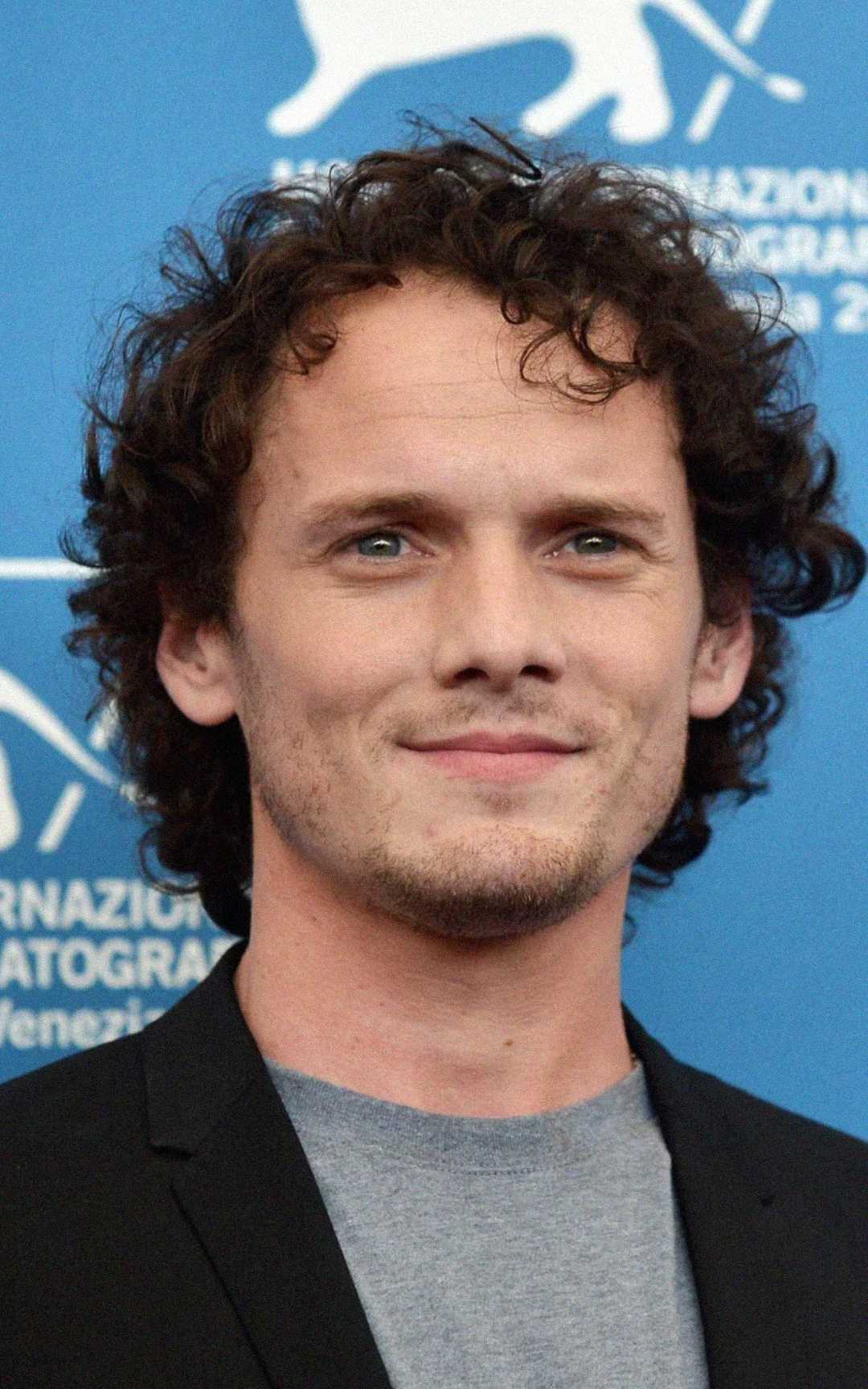 A man with curly brown hair and blue eyes is smiling slightly. He is wearing a gray shirt and a black blazer, standing in front of a blue background with white text and a lion logo.