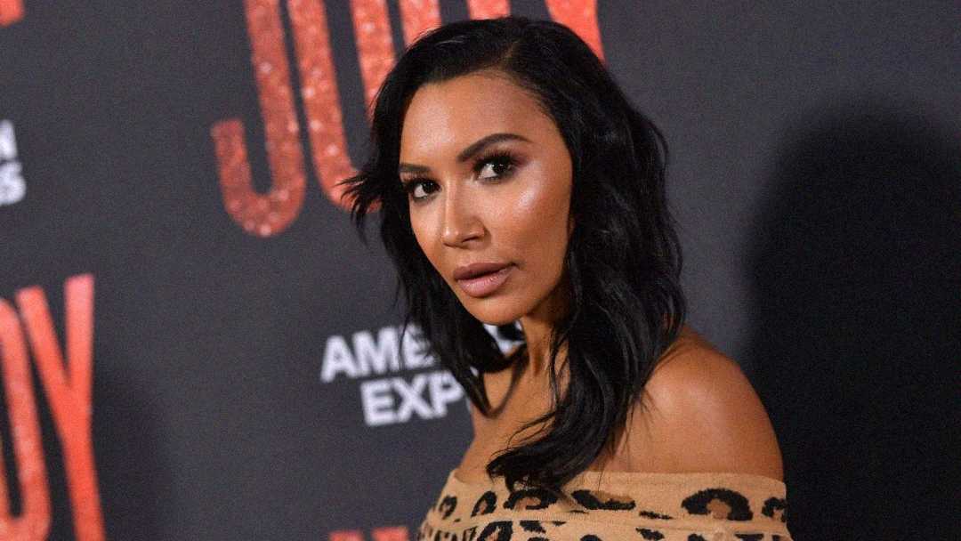 A woman with long, wavy dark hair wearing an off-the-shoulder, leopard-print top poses in front of a backdrop with red glittery text and an American Express logo.