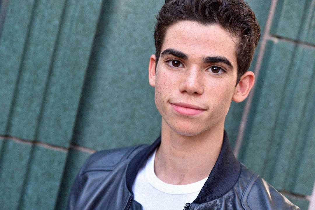 A young person with short, curly dark hair wearing a black leather jacket and white shirt stands in front of a green tiled wall, looking at the camera with a slight smile.