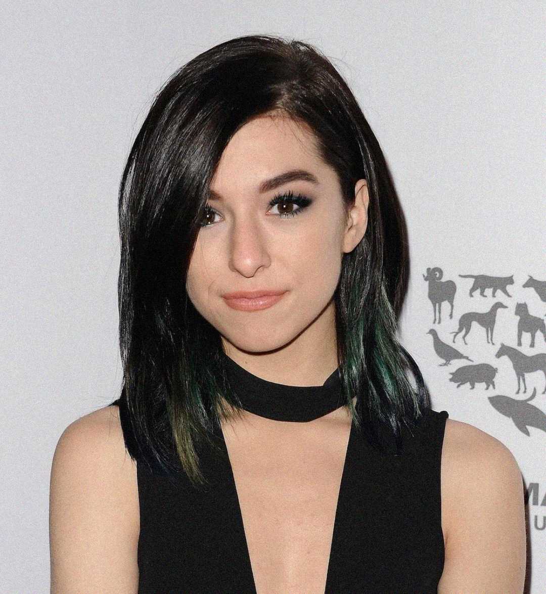 A woman with straight, dark shoulder-length hair and subtle makeup wears a sleeveless black dress with a choker neckline, standing in front of a light gray background with animal silhouettes.