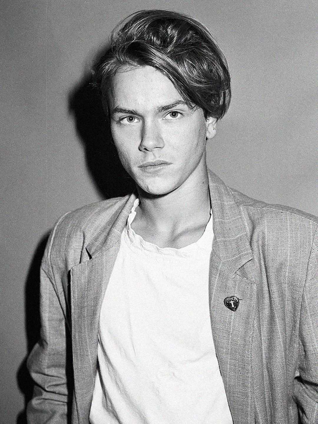Black and white photo of a young person with medium-length hair wearing a light-colored blazer over a white t-shirt, looking seriously at the camera against a plain background.
