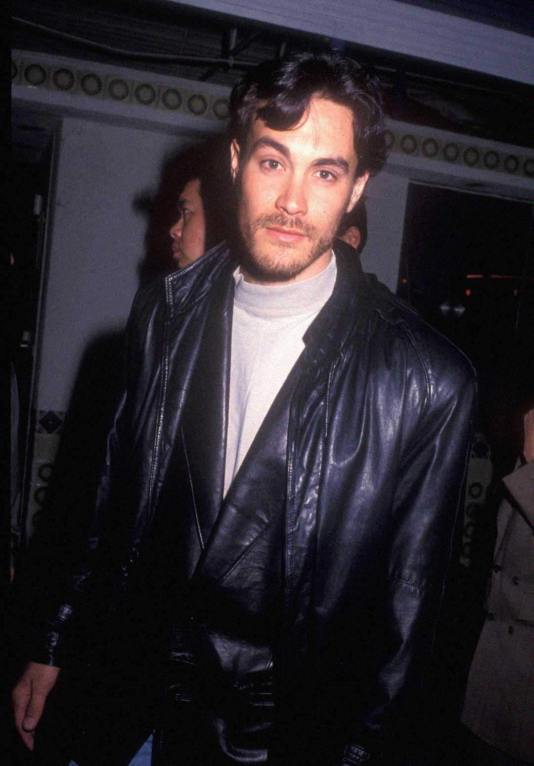 A man with wavy dark hair and facial hair wears a black leather jacket over a light-colored shirt, standing indoors at an event with other people visible in the background.