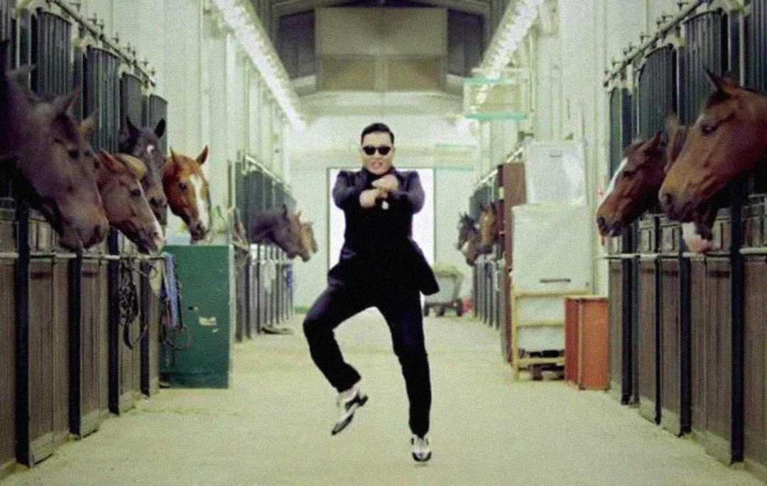 A person in sunglasses and a suit dances in the middle of a stable corridor, with several horses watching from their stalls on both sides.