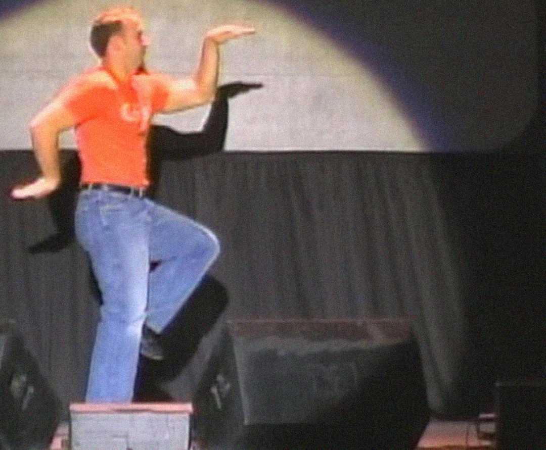 A person in a red shirt and blue jeans strikes a dramatic pose on stage, balancing on one leg with both arms bent, while performing under a spotlight.