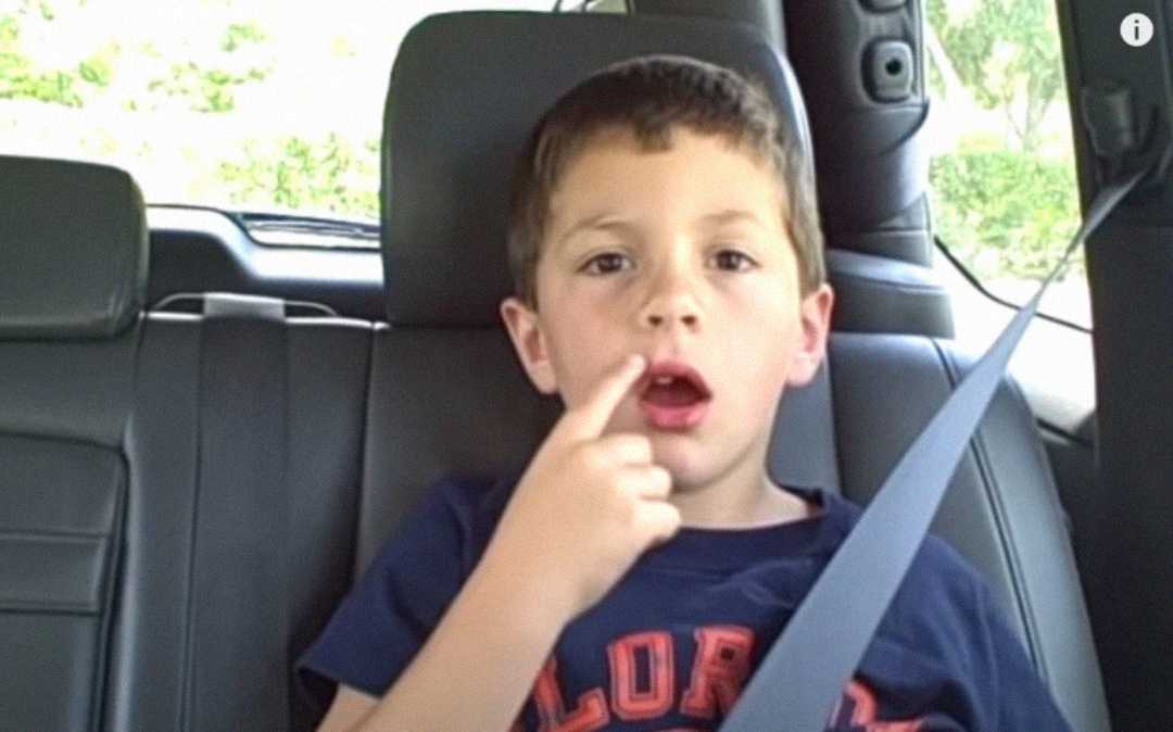 A young boy sits in the back seat of a car, wearing a seatbelt and a dark blue shirt. He has his mouth slightly open and is touching his cheek with his finger, looking slightly surprised.