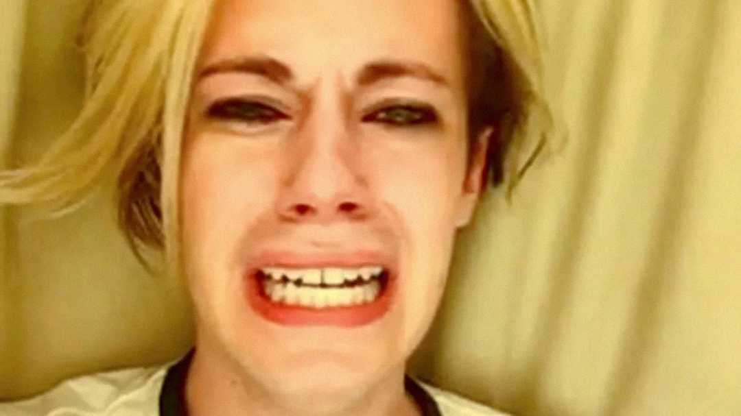 A person with blonde hair and heavy eye makeup is crying with an anguished expression, mouth open and teeth showing. The background is a plain beige curtain.