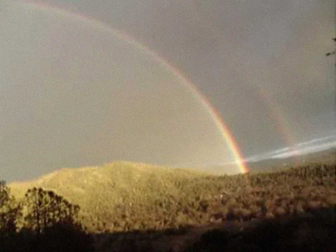A double rainbow arcs across a cloudy sky above sunlit, tree-covered hills, with the landscape bathed in warm, golden light.