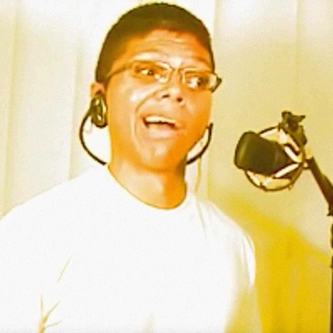 A person wearing glasses and a headset stands in front of a microphone, appearing to sing or speak. The background is softly lit with light-colored curtains.