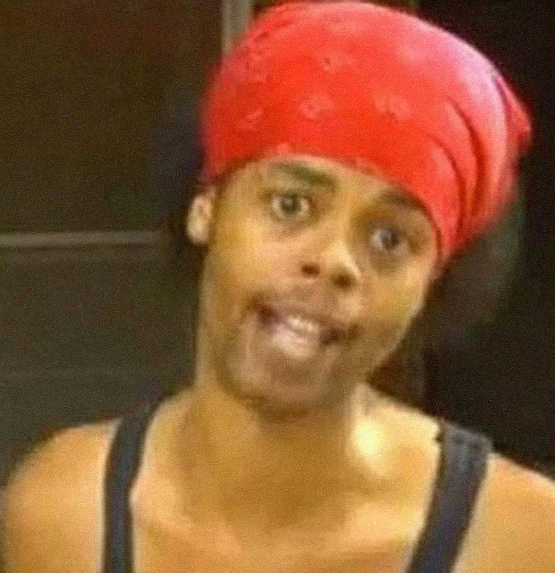 A person wearing a red bandana and black tank top looks directly at the camera with a slightly open mouth, appearing to speak, set against an indoor background.