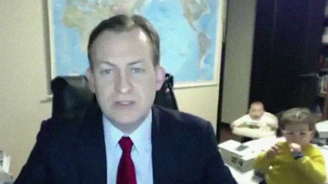 A man in a suit speaks on a video call in an office, with a world map on the wall behind him. Two young children play in the background, one in yellow and the other in white.