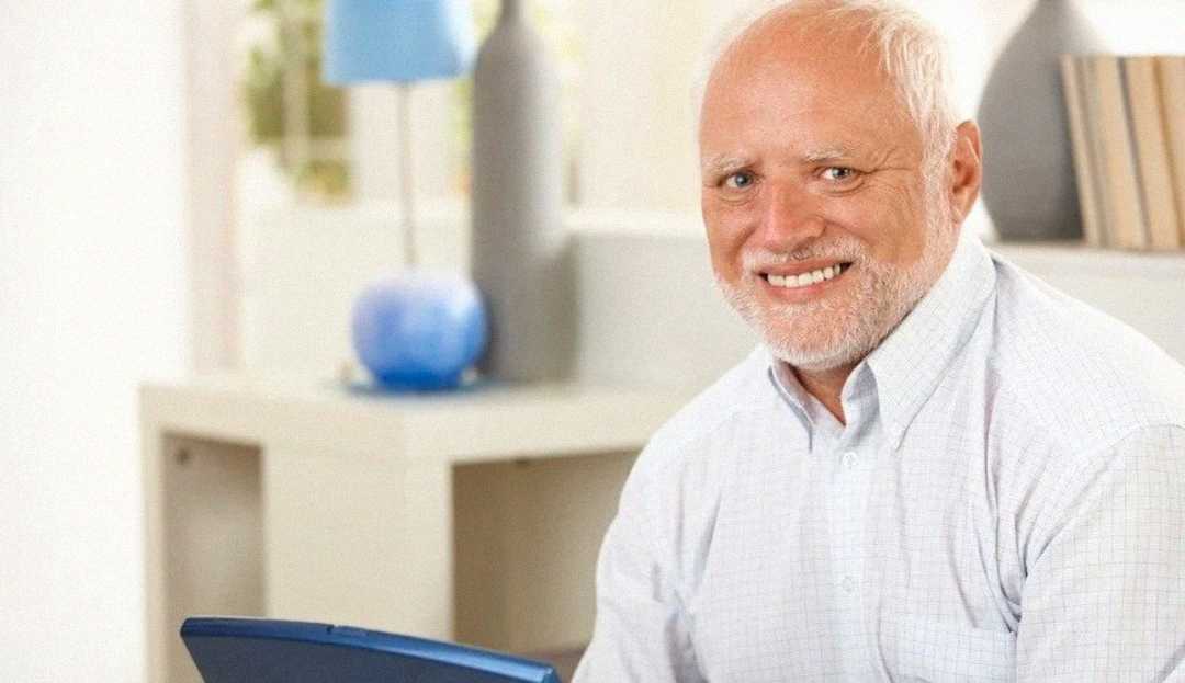 An older man with white hair and a beard is sitting indoors, smiling while holding a blue laptop. He is wearing a light-colored, button-up shirt. The background features a lamp, books, and a decorative vase.