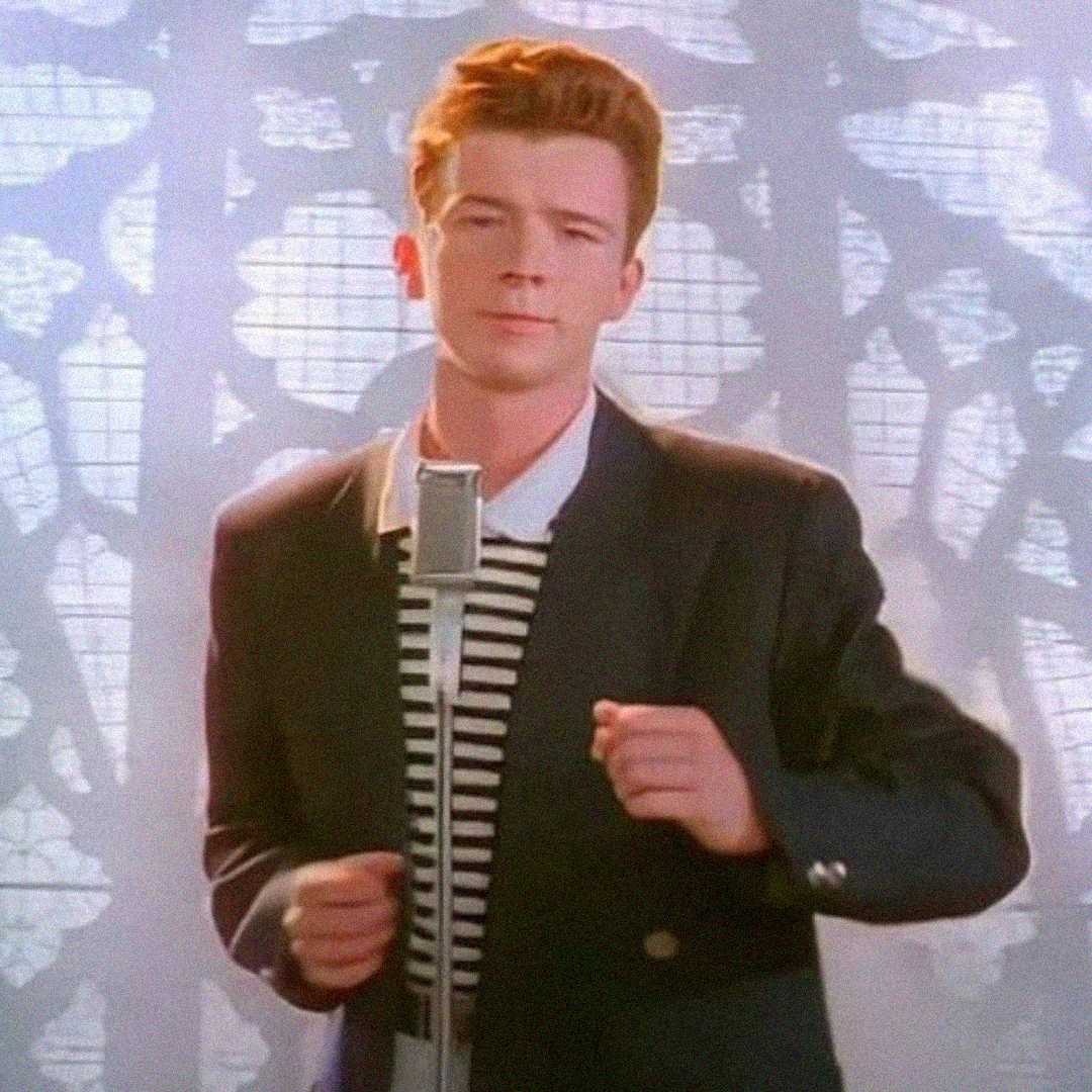 A man with short, reddish-brown hair stands in front of a vintage microphone, wearing a black blazer over a black and white striped shirt, with a patterned background behind him.