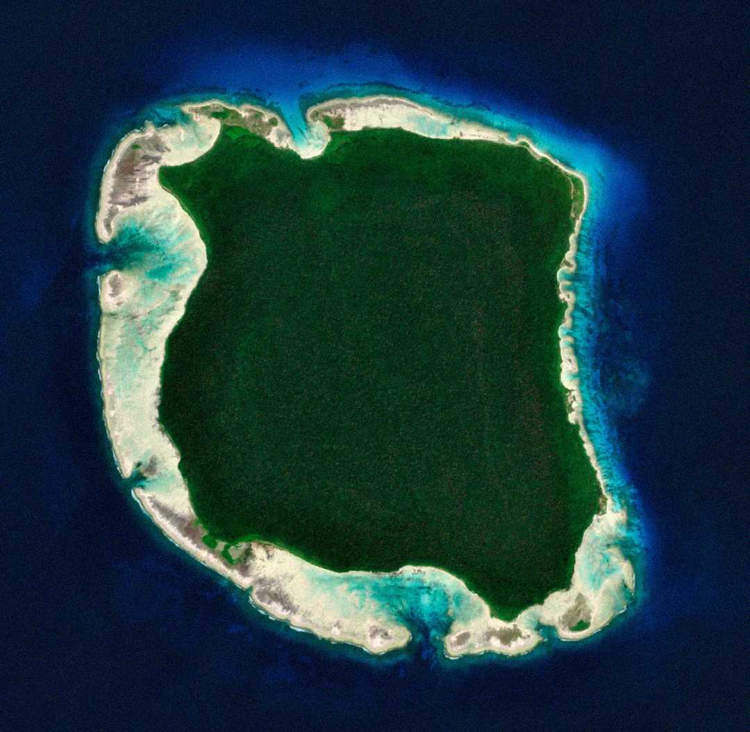 Aerial view of a densely forested, roughly square-shaped island surrounded by a narrow strip of sandy beach, clear turquoise waters, and deep blue ocean. No visible signs of human activity or structures.