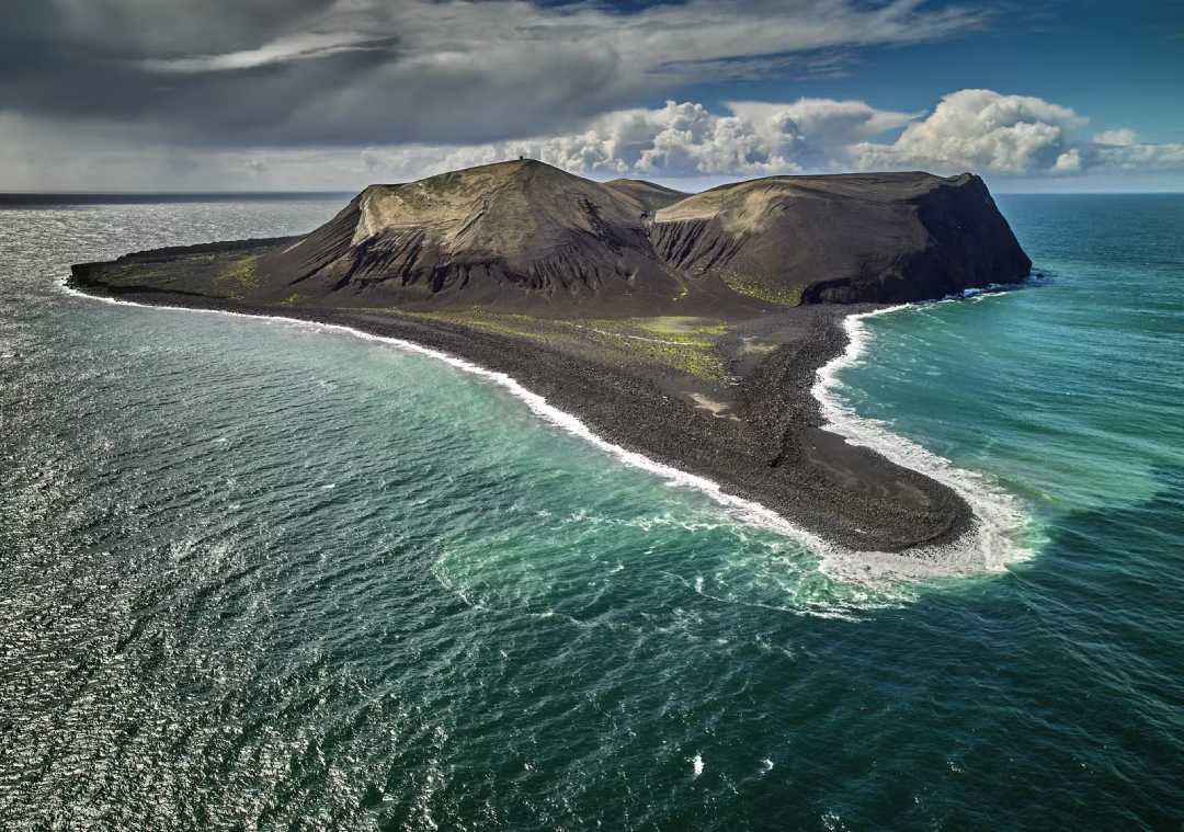 A rugged volcanic island rises from the ocean, featuring steep, rocky slopes, sparse vegetation, and surrounded by turquoise and deep blue waters under a partly cloudy sky.