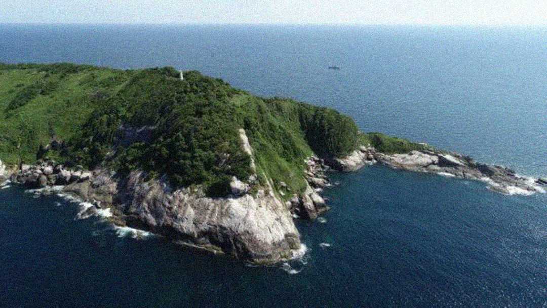 A rocky, green-covered peninsula juts into the deep blue ocean under a clear sky, with gentle waves touching the shoreline and a boat visible in the distance.