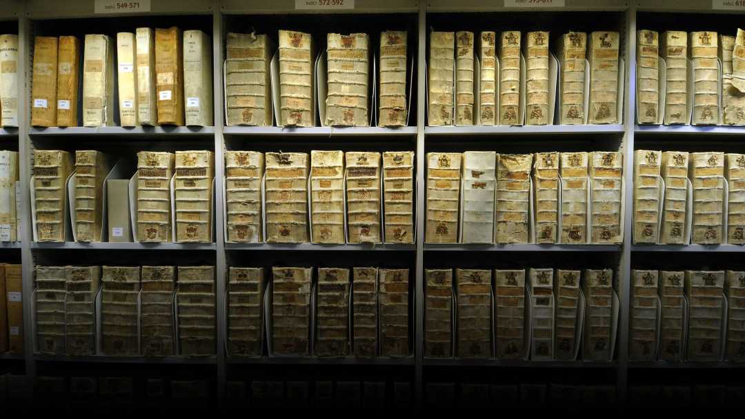 Rows of old, worn books and documents with faded covers and labels are neatly arranged on shelves, suggesting an archive or historical library collection. The lighting highlights the aged appearance of the materials.