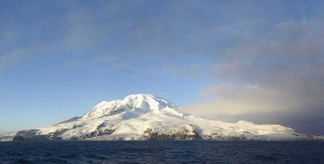 Snow-covered volcanic island rises from the ocean under a clear blue sky, with some clouds to the right and rocky, icy coastline in the foreground.