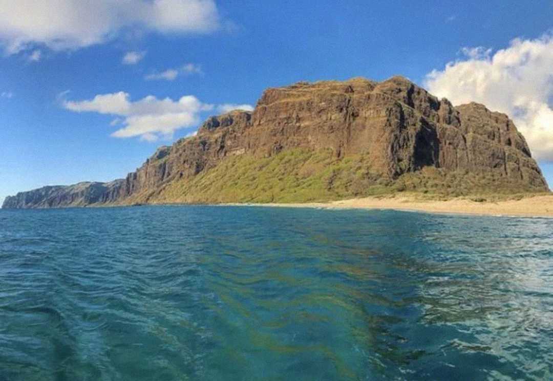 A rugged, rocky cliff rises from a sandy beach, surrounded by clear blue ocean water under a mostly sunny sky with some scattered clouds.