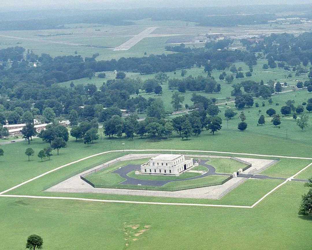 Aerial view of a fortified building surrounded by a fence and green fields, with trees scattered around and a road leading to the entrance; additional buildings and a runway are visible in the background.