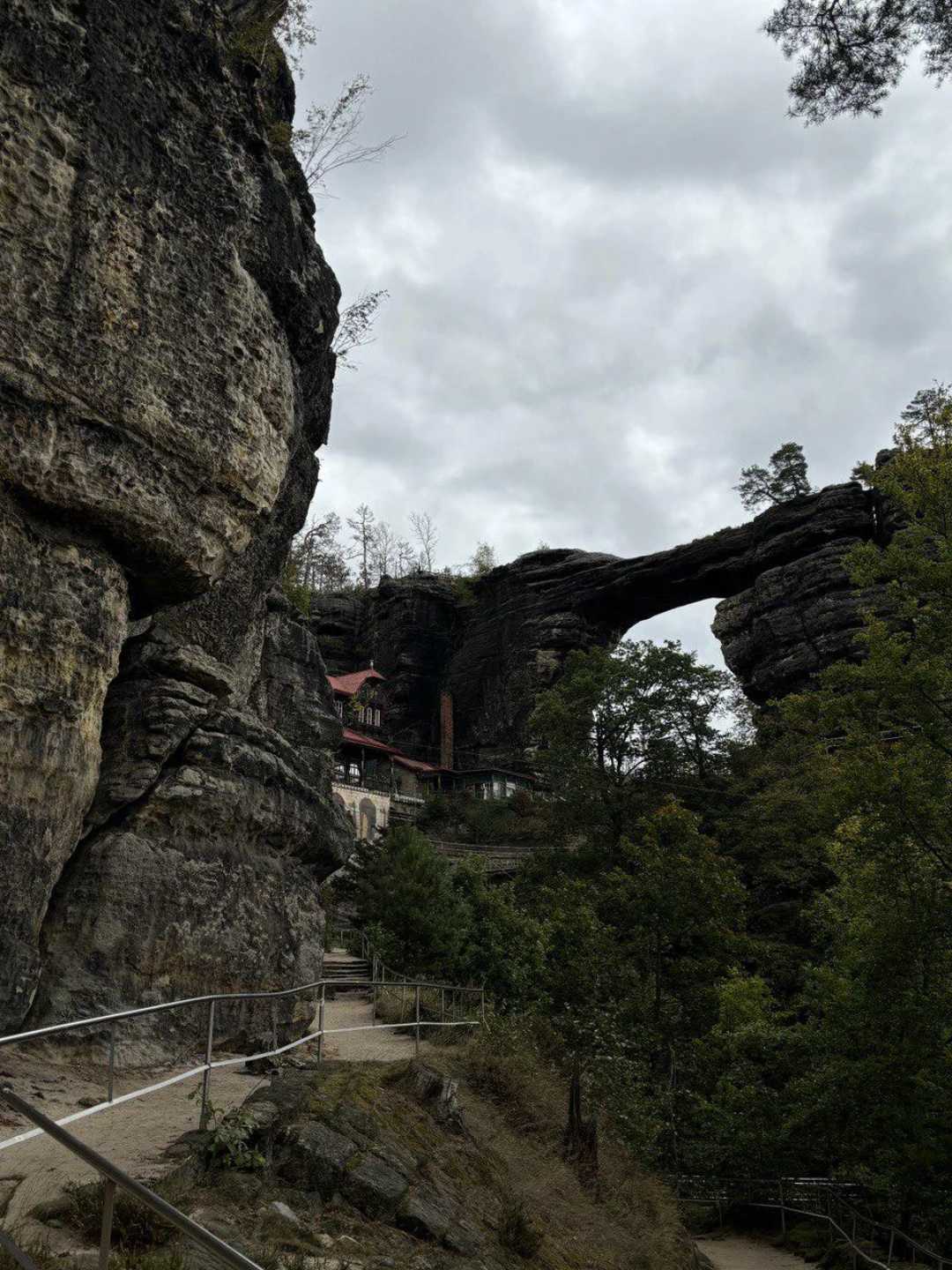 A rocky trail with railings winds past large stone cliffs and trees, leading to a natural rock arch formation. A building with a red roof is nestled beneath the arch under a cloudy sky.
