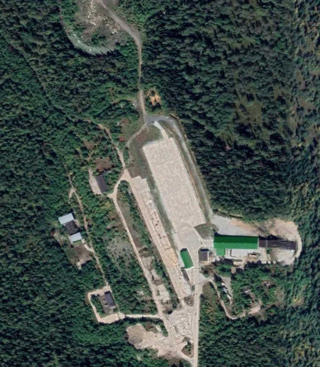 A satellite image showing a rectangular concrete area surrounded by dense green forest, with several small buildings and dirt roads nearby.