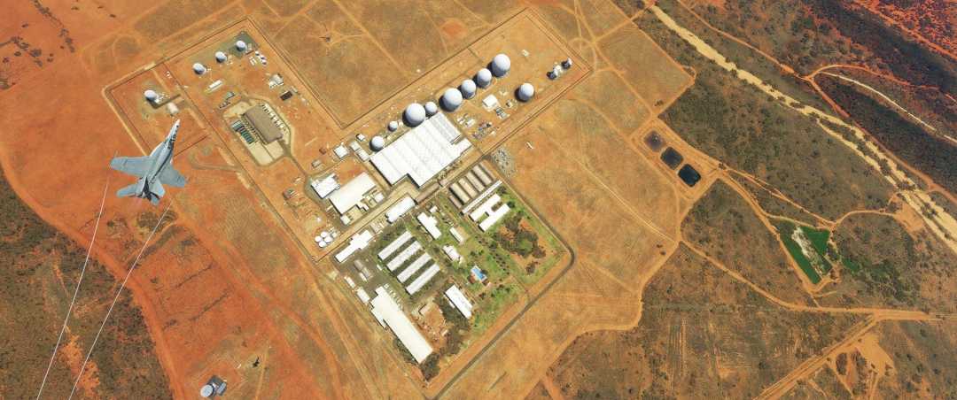 Aerial view of a fenced desert facility with several white domes, buildings, and roads. A military jet flies nearby with its shadow visible on the red earth surrounding the compound.