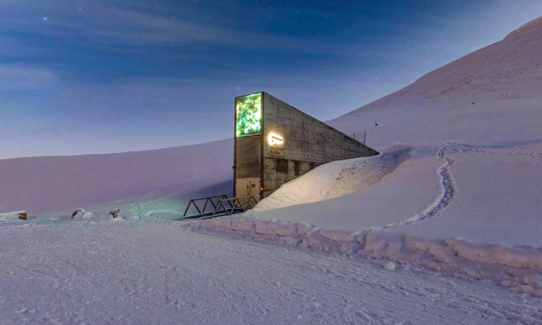 A modern, concrete structure partly buried in snowy terrain, illuminated by a green-lit sign near its entrance; mountains and a clear sky are in the background.