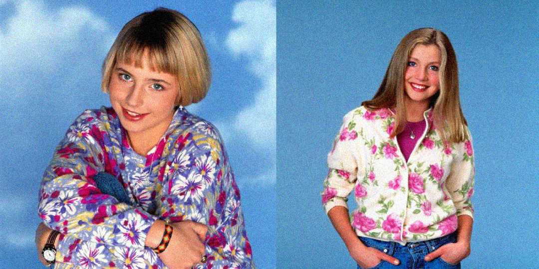 Two girls pose in front of a blue background. The girl on the left has a short blonde bob and wears a floral shirt. The girl on the right has long blonde hair and wears a white sweater with pink flowers and blue jeans.