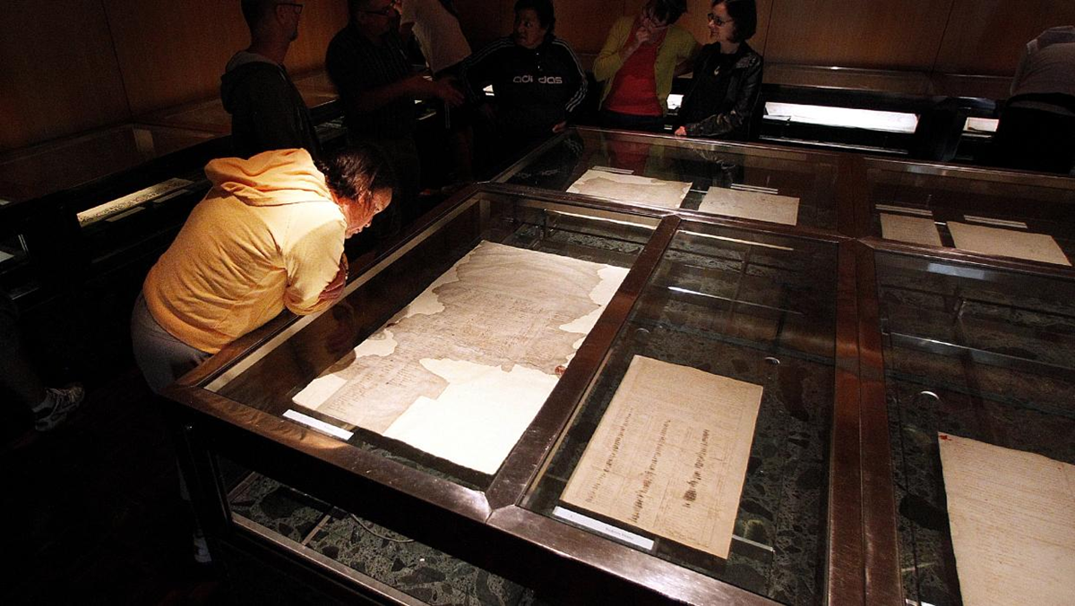 A person in a yellow hoodie closely examines historic documents displayed in glass cases at a dimly lit museum, while other visitors observe in the background.