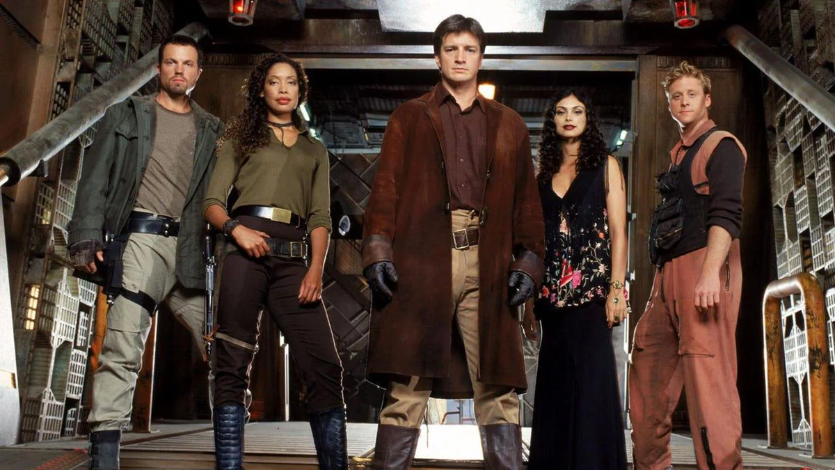 Five people in varied futuristic outfits stand confidently in a metal spaceship corridor, facing forward. The central figure wears a long brown coat; others wear utility gear or a black dress. The setting has an industrial, sci-fi look.